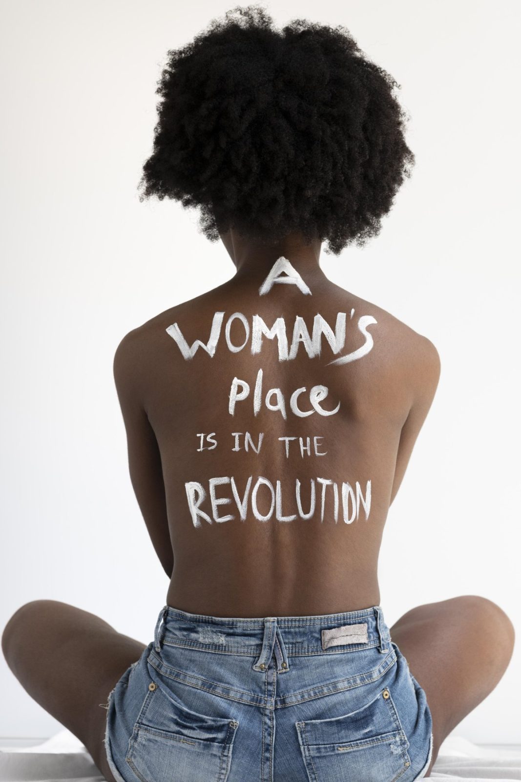 Dos d’une femme assise, portant peinte en blanc la phrase « A woman’s place is in the revolution ».